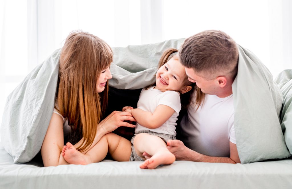 California family in sheets of their bed, experiencing the benefits of regular HVAC maintenance: laughter, comfort, and energy savings